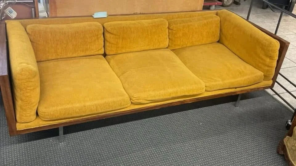 a mustard-yellow, mid-century modern style couch with thick cushions and wooden frame, photographed in what appears to be a…