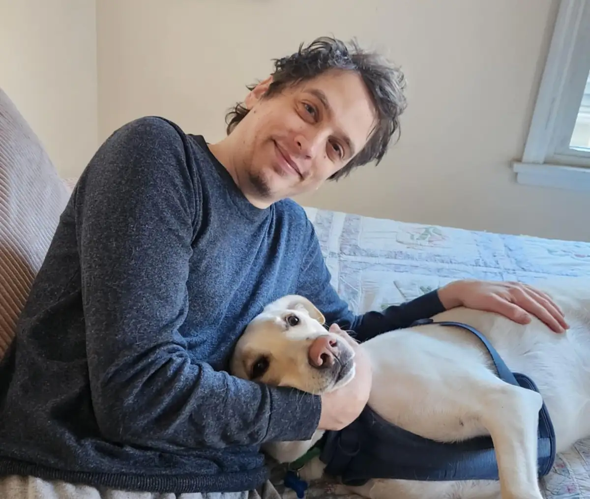 A man with dark, curly hair and a slight smile is sitting on a bed with a light-colored Labrador Retriever. The dog is lying…