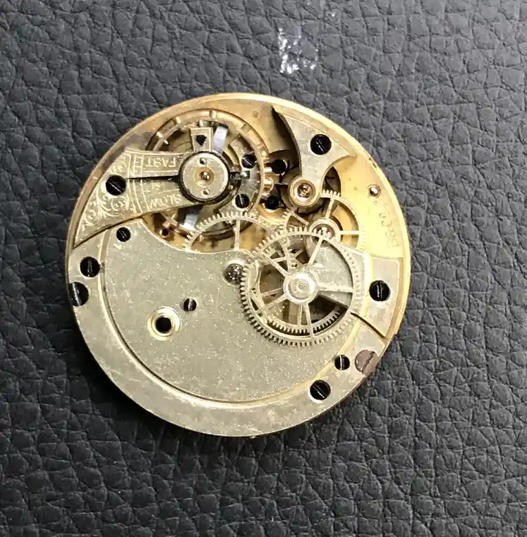 A close-up photo of a golden mechanical watch movement on a dark textured surface, with an engraved lever clearly marked…