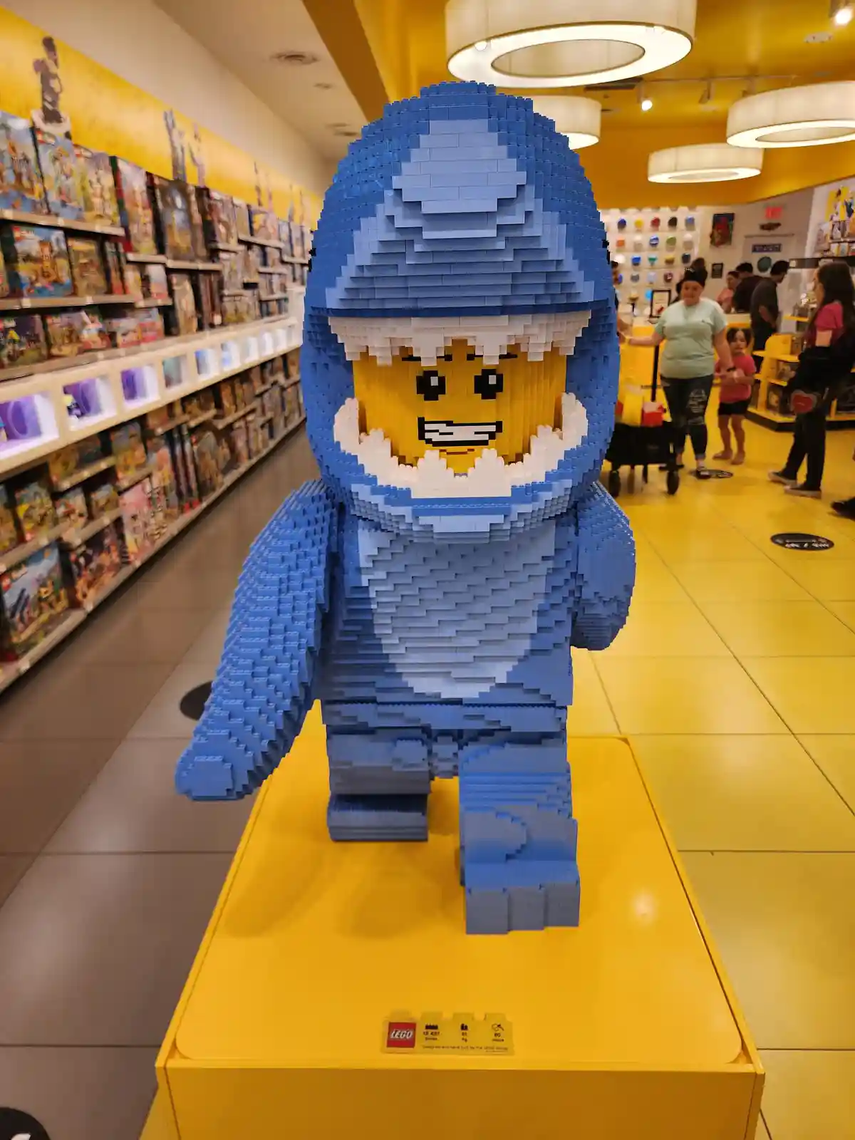 A giant LEGO minifigure statue, constructed from blue and white bricks, stands on a yellow pedestal inside a brightly lit…