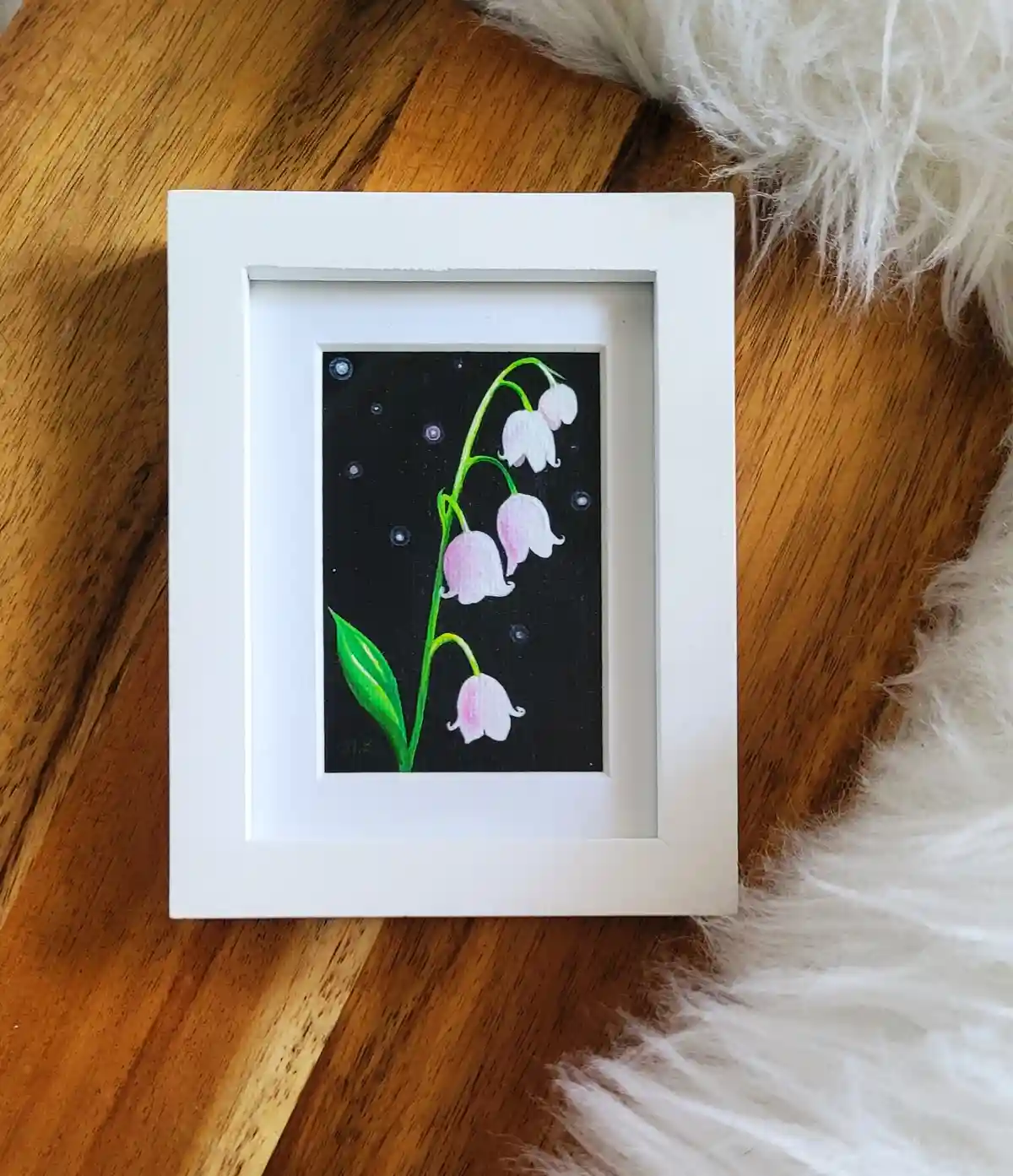 A small white-framed painting depicts a lily of the valley plant with pink bell-shaped flowers against a black background…