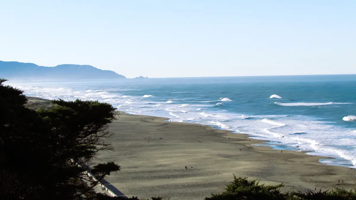 A wide-angle photograph of a scenic beach with rolling ocean waves, distant mountains, and sparse people on the sand. The…