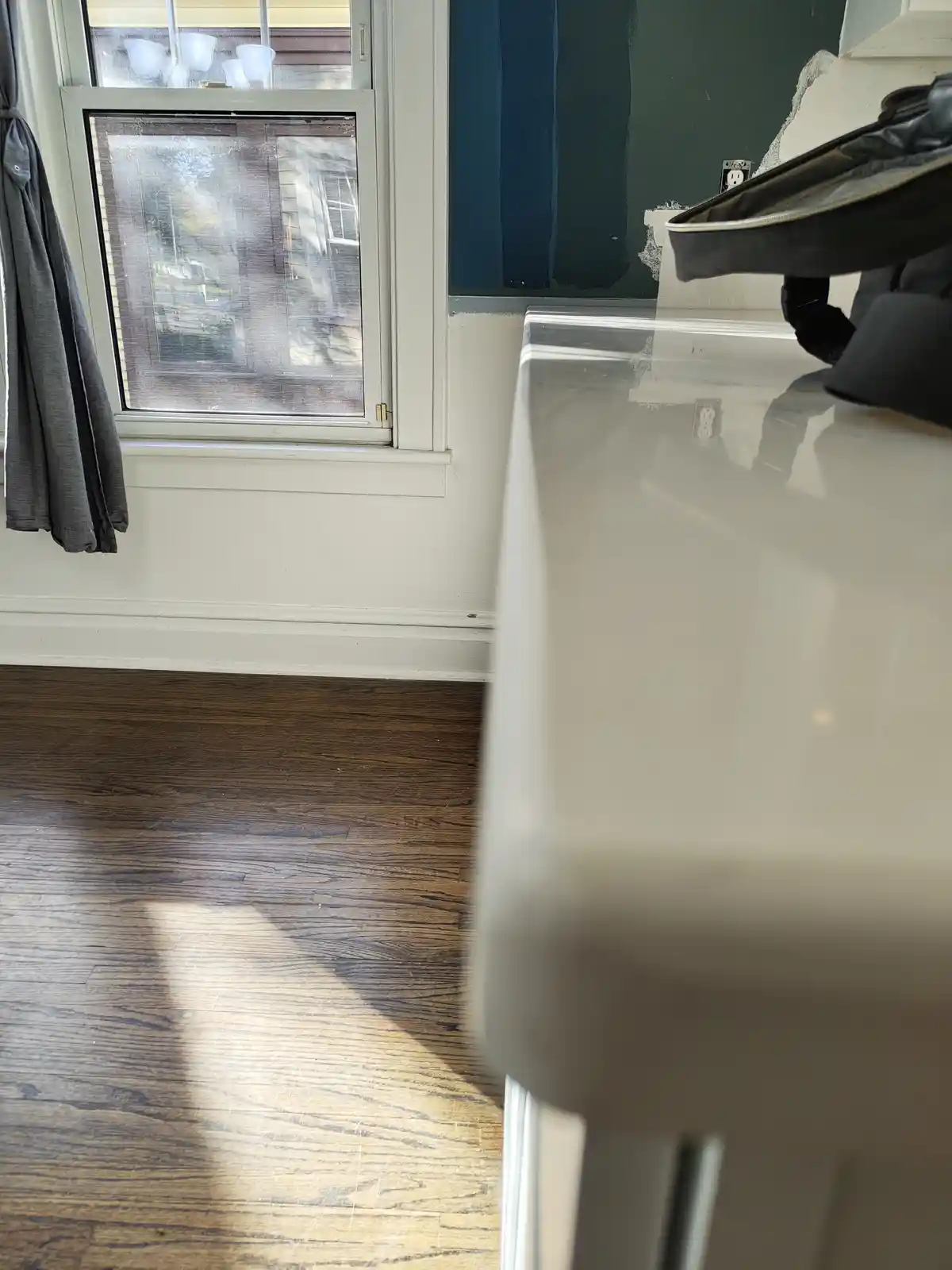 A photo shows a partially painted kitchen wall next to a window with a grey curtain, a white countertop with a black bag on…