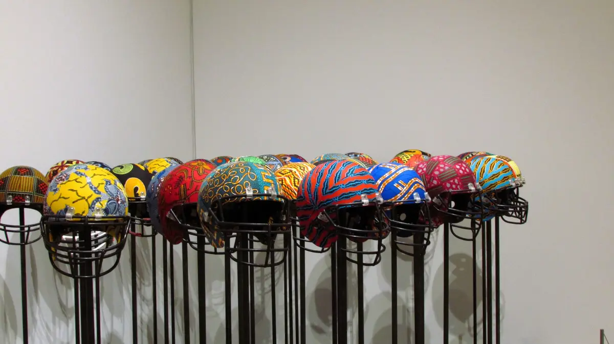 A row of American football helmets, each decorated with vibrant, colorful African wax print patterns, are displayed on black…