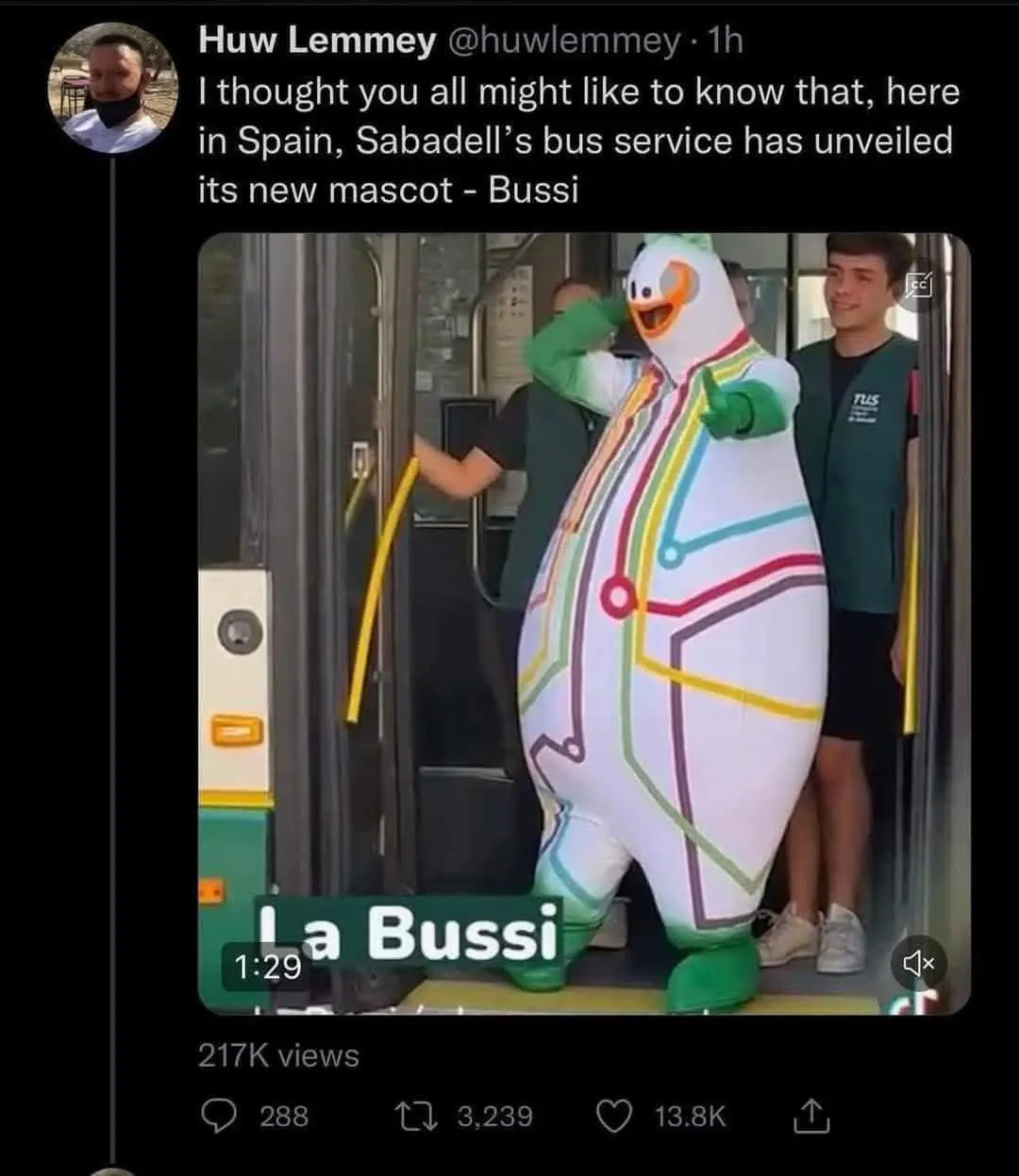 A person in a large, white, inflatable duck mascot costume with colorful lines resembling a bus route stands in the doorway…