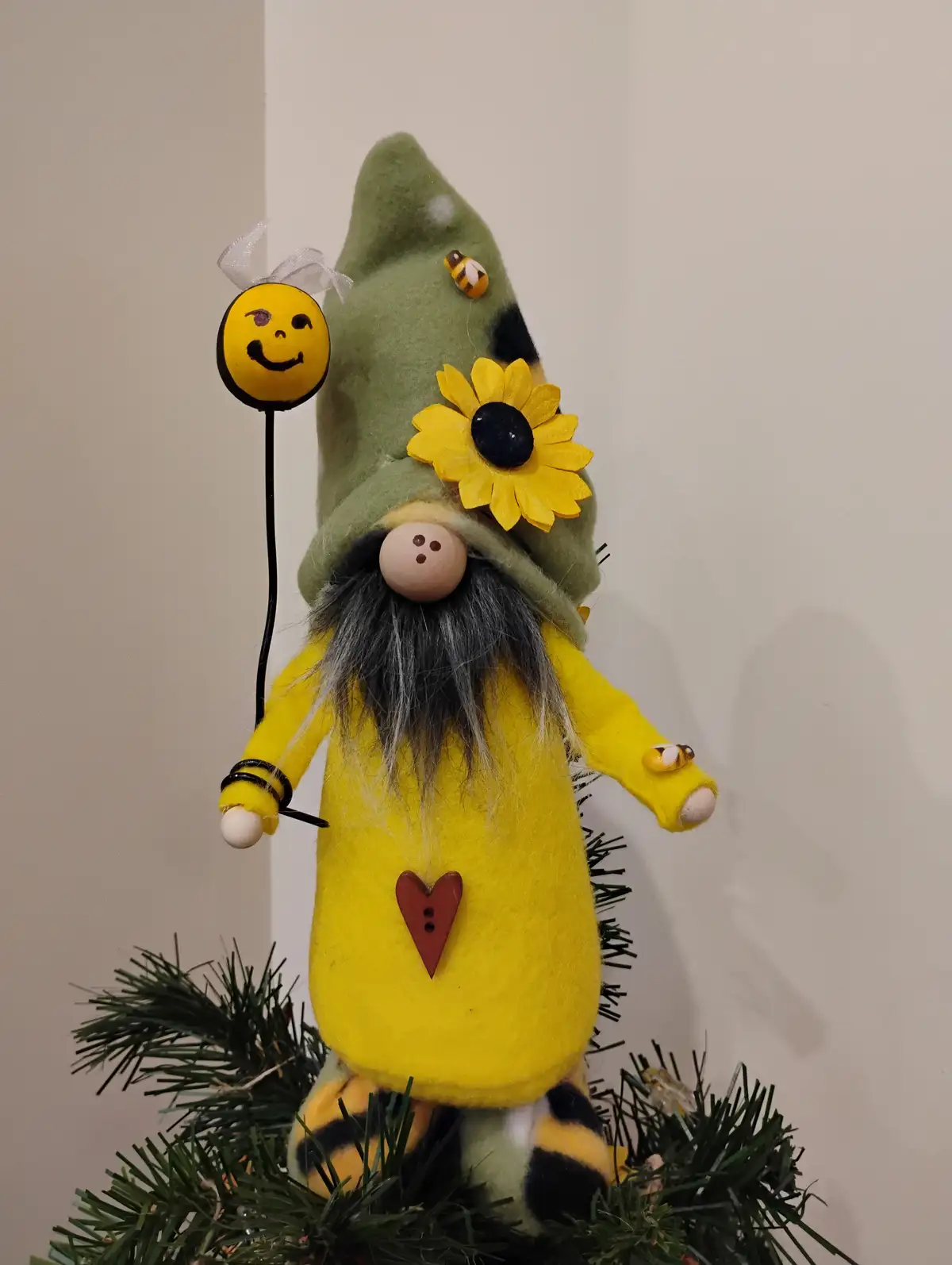 A handmade felt gnome wearing a yellow outfit and green hat, standing on a Christmas tree with a sunflower decoration and…