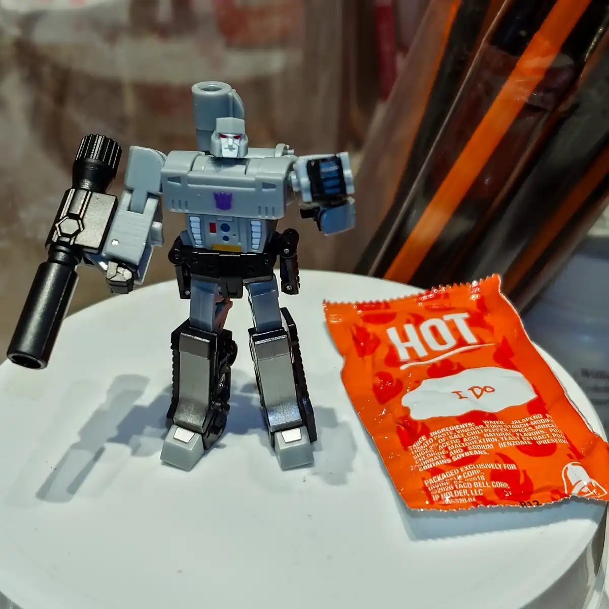 A gray and black Transformers toy robot stands next to an orange Taco Bell hot sauce packet. The packet has 'HOT' printed in…