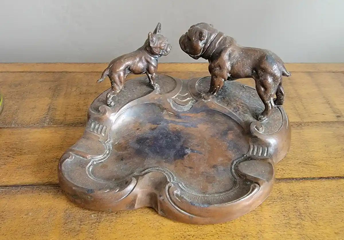 A vintage bronze ashtray features two small dog statues, one resembling a French bulldog and the other a bulldog, standing…
