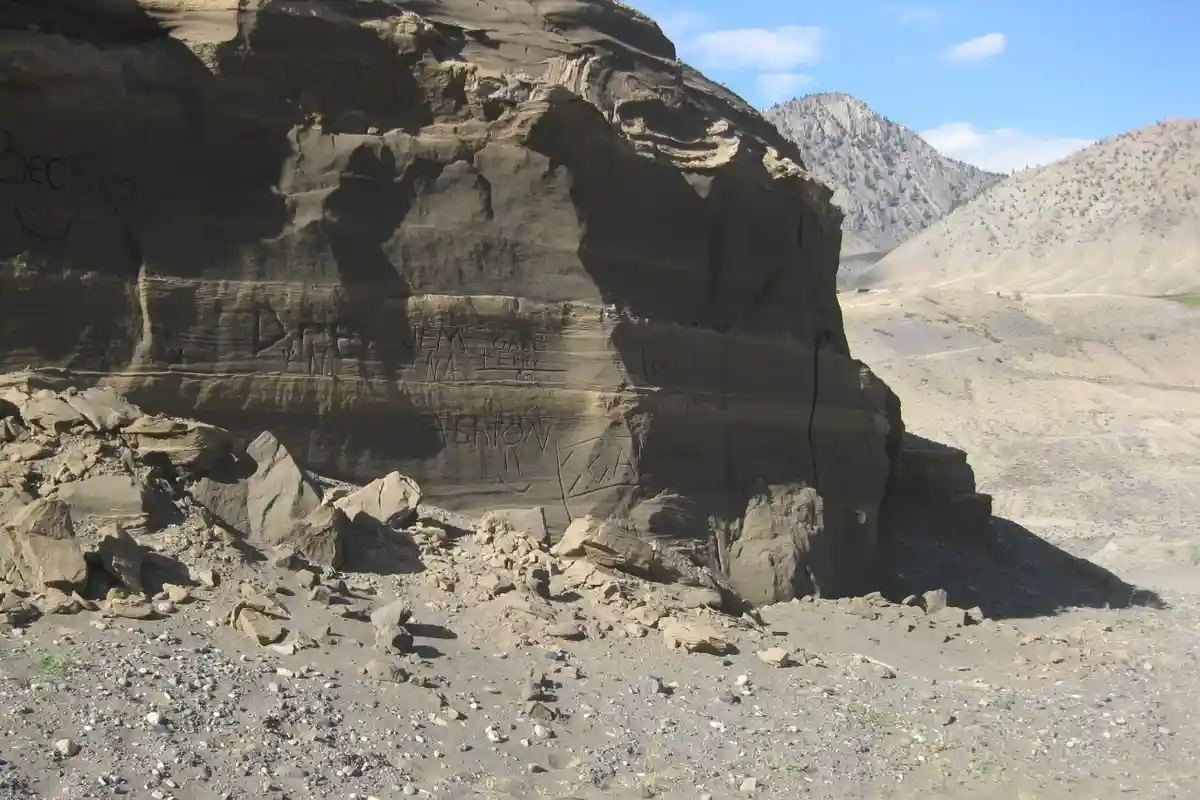 A large, sunlit rock face in a desert canyon is covered with numerous carved names and initials, including 'Drew', 'Jem'…
