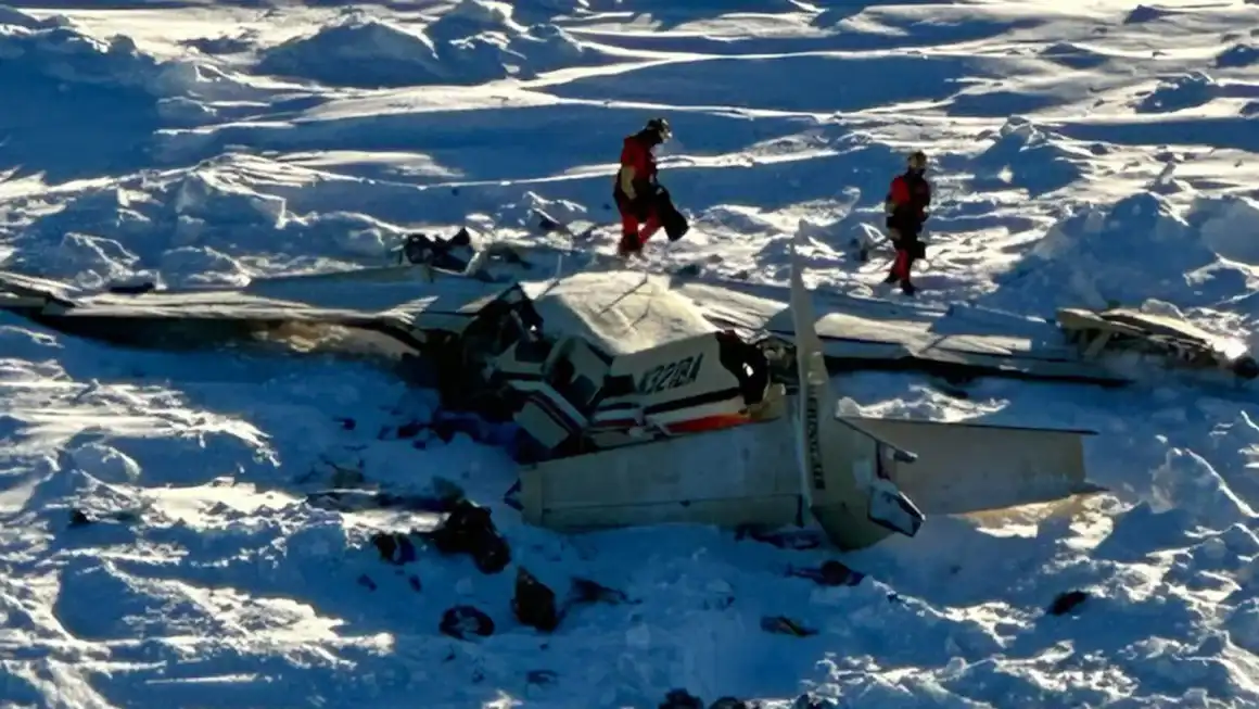 A crashed airplane lies in a snowy, mountainous landscape, with two rescuers in red gear walking nearby. The text overlaid…