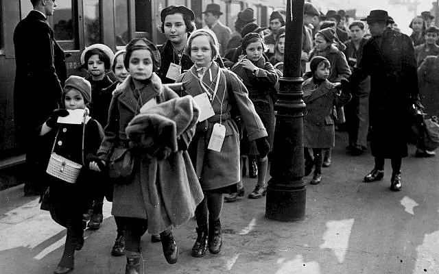 A black-and-white historical photograph shows a group of children, some wearing coats and hats, walking on a platform near a…