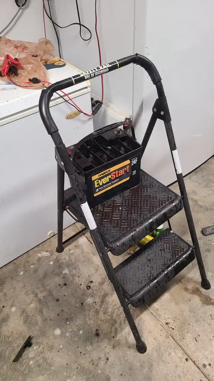 A black, two-step metal step ladder sits in a garage, with a black EverStart car battery placed on its top step. Red and…