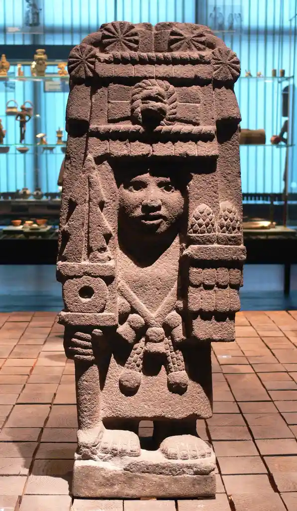 A large, intricately carved ancient stone statue of a human figure with elaborate headdress and ceremonial attire stands in…