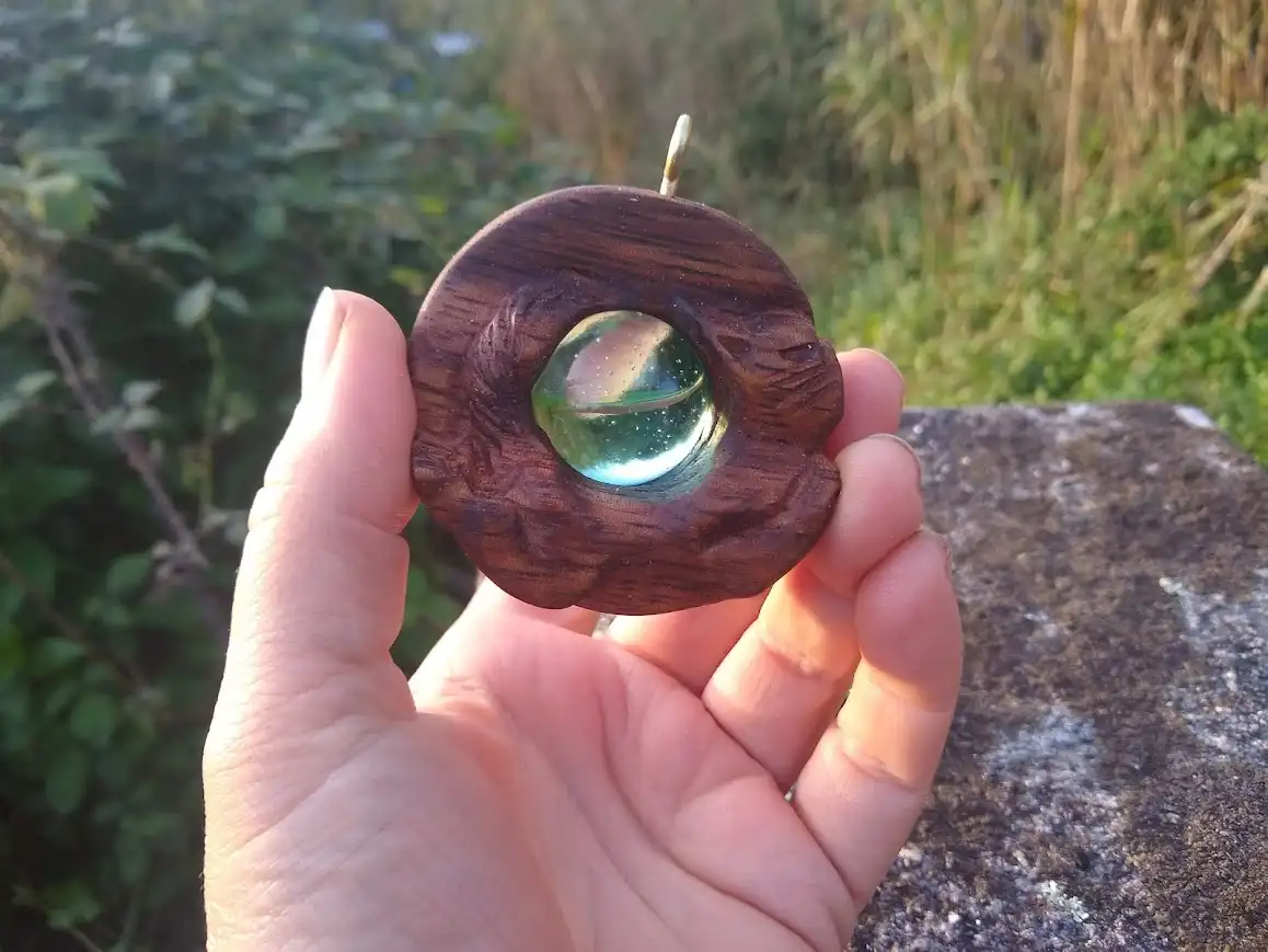 A hand holds a dark wooden circular pendant with a glass orb embedded in its center, set against a blurred natural…