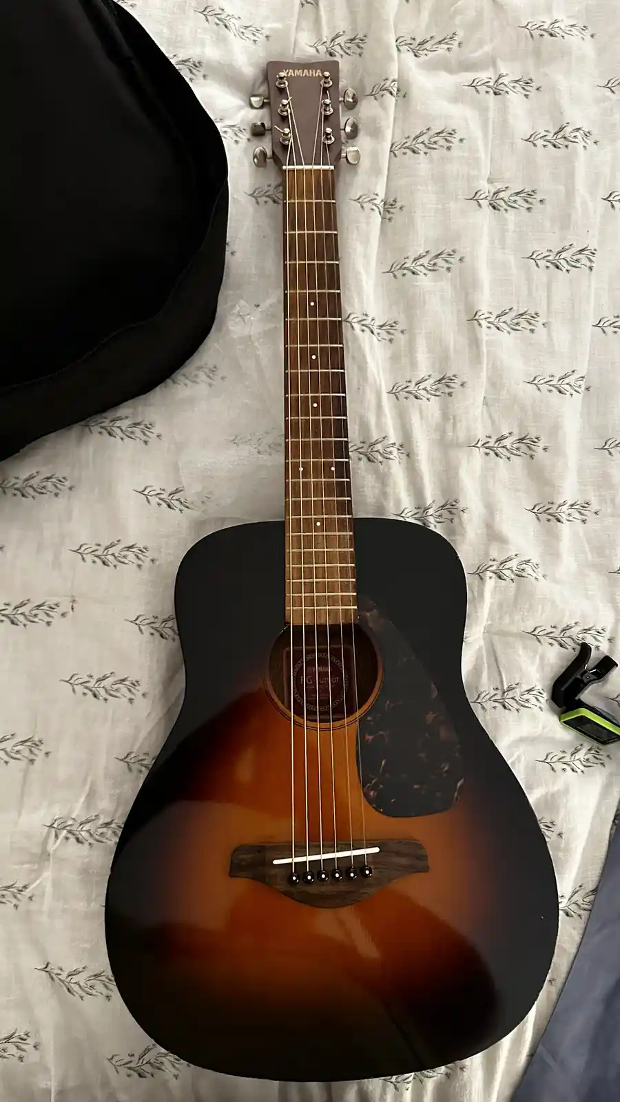 A Yamaha acoustic guitar with a sunburst finish lies on a white fabric with a leaf pattern, next to a black guitar case and…