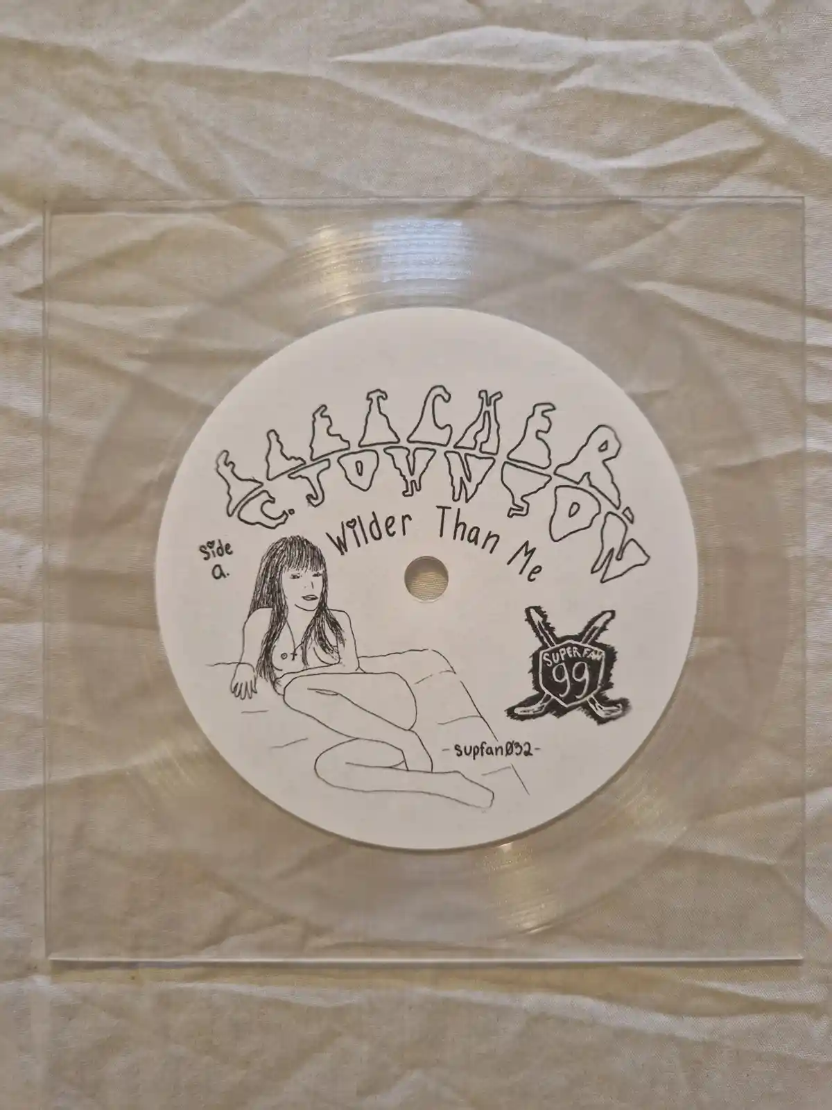 A close-up photo of a handmade, transparent vinyl record label inside a clear sleeve, resting on a beige fabric. The label…