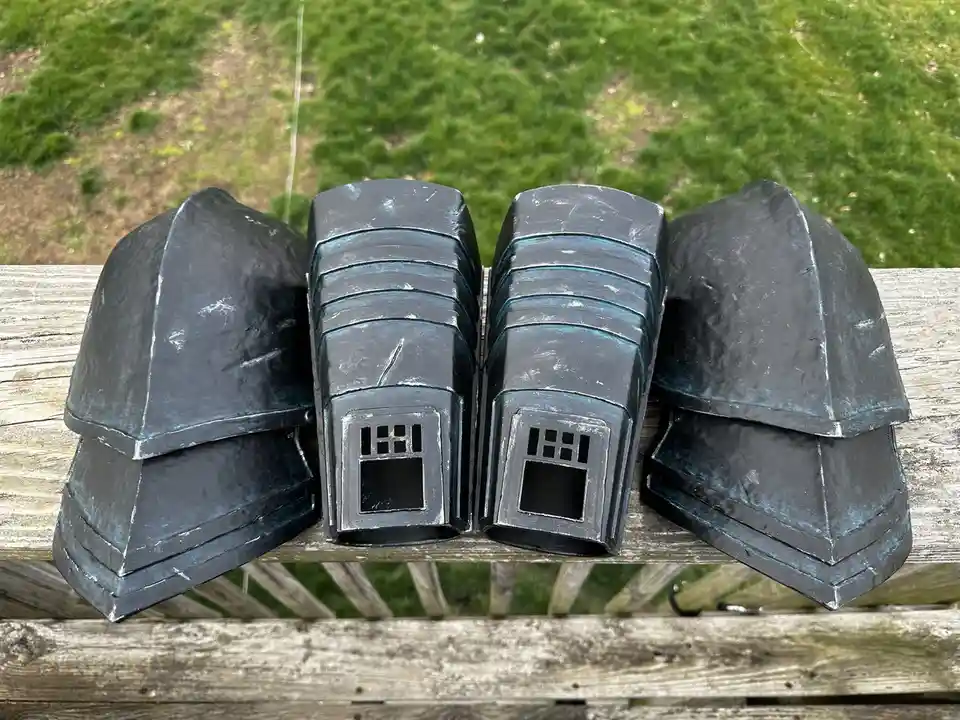 Four black, hand-painted, house-shaped objects with windows sit on a weathered wooden bench outdoors, with grass in the…