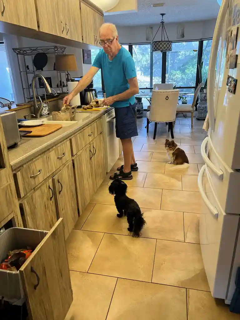 An older man in a blue shirt and shorts stands at a kitchen sink, holding a piece of raw meat, looking concerned. Two small…