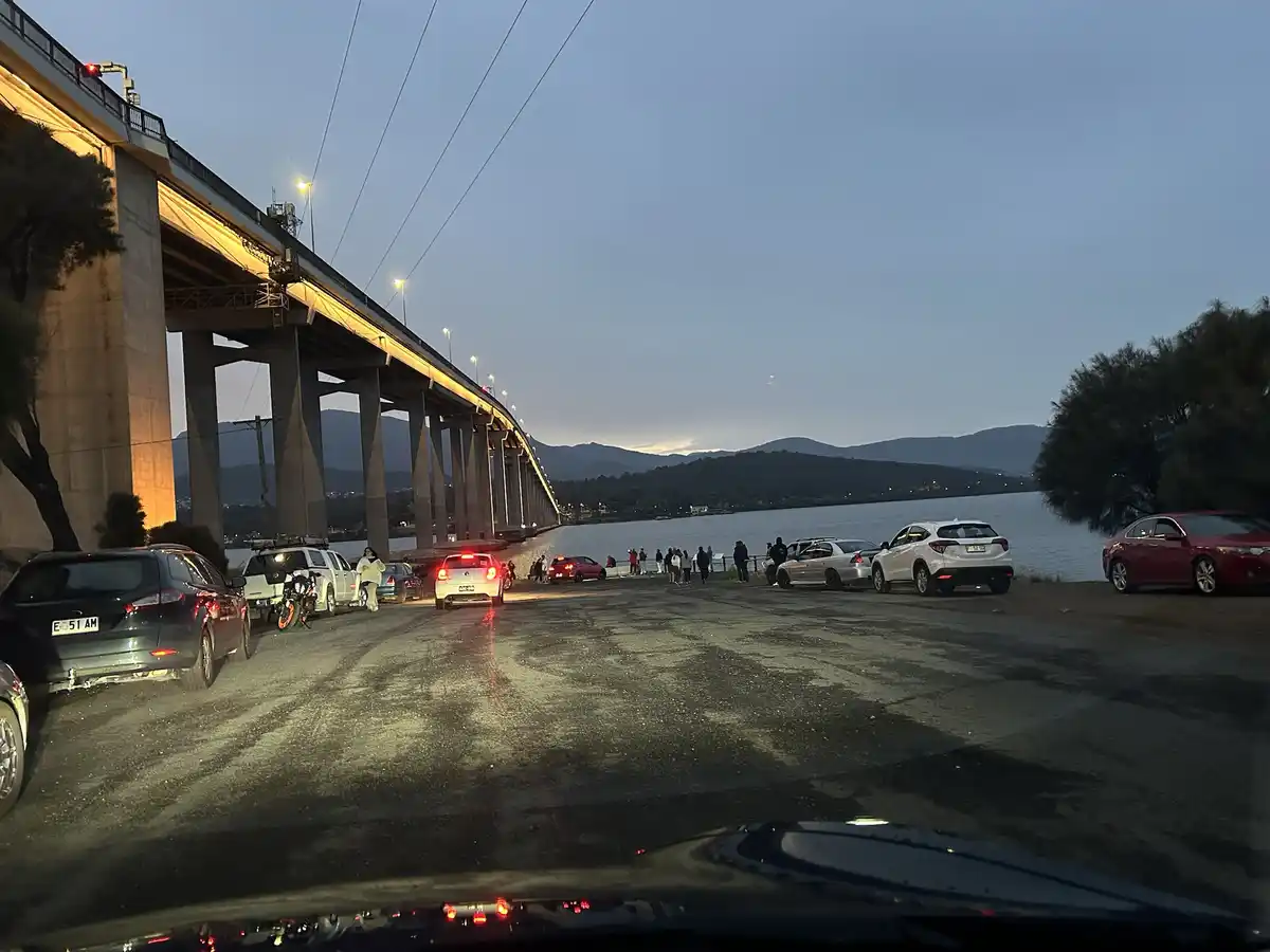 A photo taken from inside a car shows a line of parked vehicles and people gathered near a large bridge at dusk, with the…