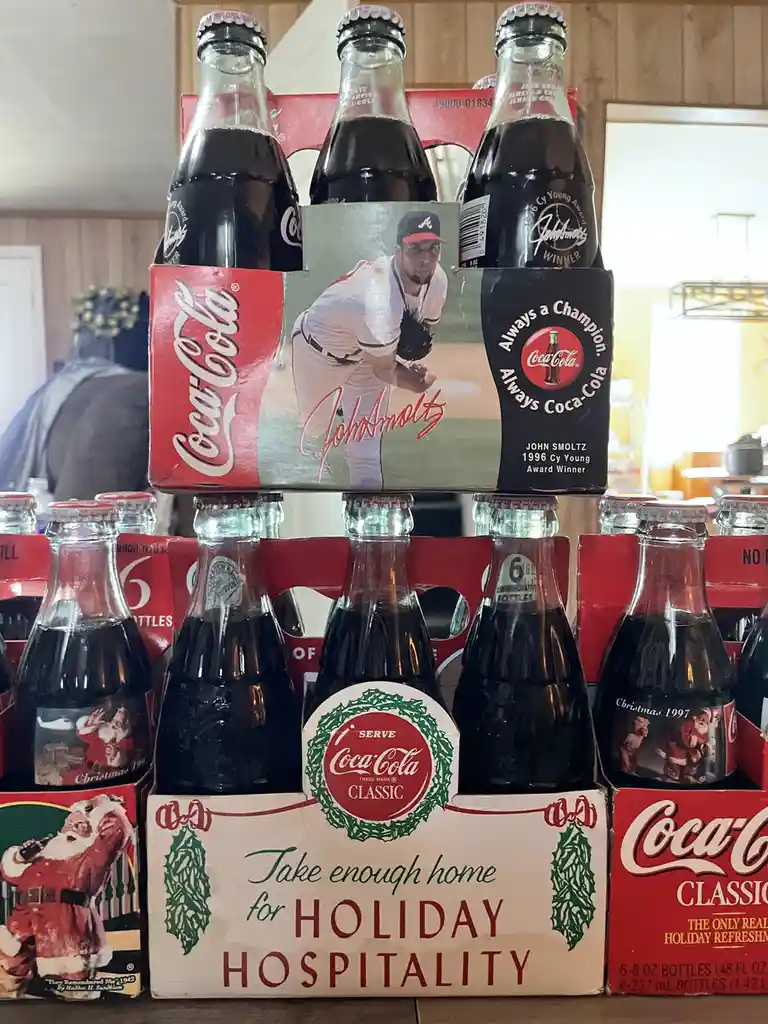 A six-pack of Coca-Cola glass bottles featuring a 1996 John Smoltz Cy Young Award commemorative label is stacked atop…