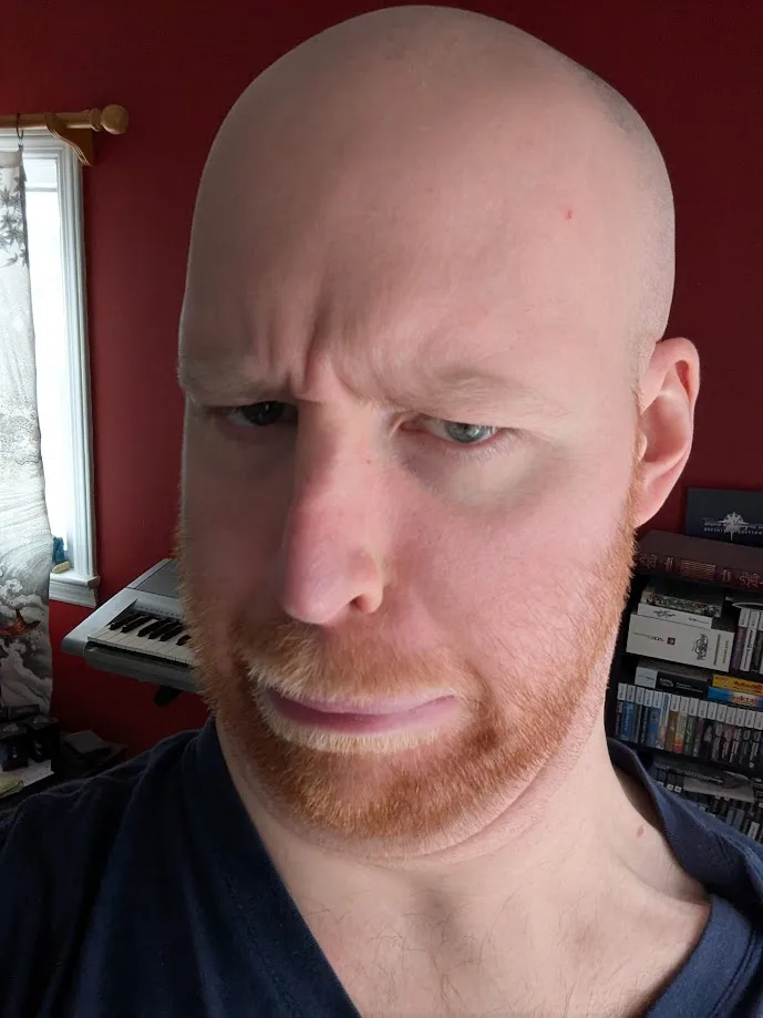 A close-up photo of a bald man with a reddish beard and mustache, looking down and to the side with a skeptical or…