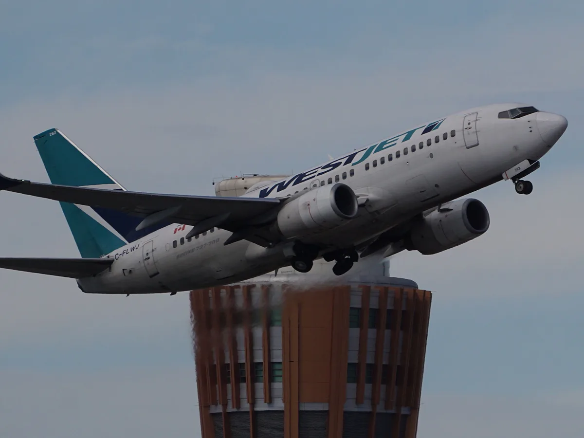 A WestJet Boeing 737-700 airplane is captured mid-air, appearing to fly directly over the top of a tall building, with its…
