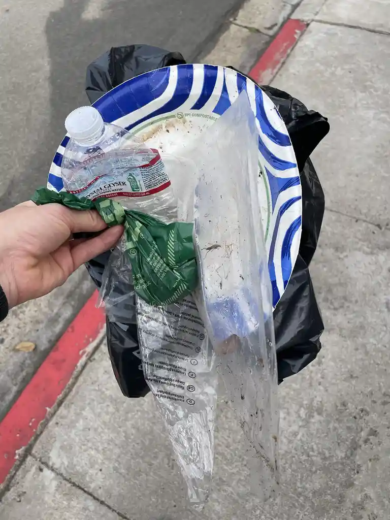 A hand holds a crumpled green plastic bag and a clear plastic bag over a black trash bag, which contains a dirty…