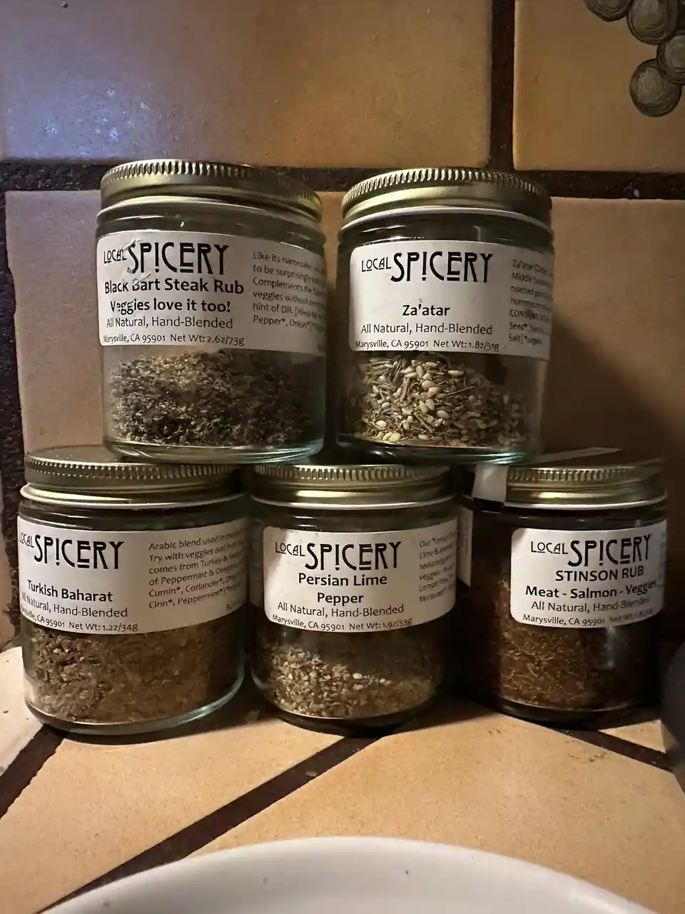 Five glass jars of 'Local Spicery' seasoning blends are stacked on a tiled countertop, each labeled with names like 'Black…