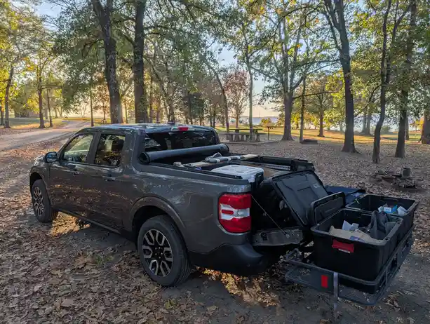 A dark gray pickup truck is parked in a wooded campground with fallen leaves on the ground. The truck bed is open and filled…