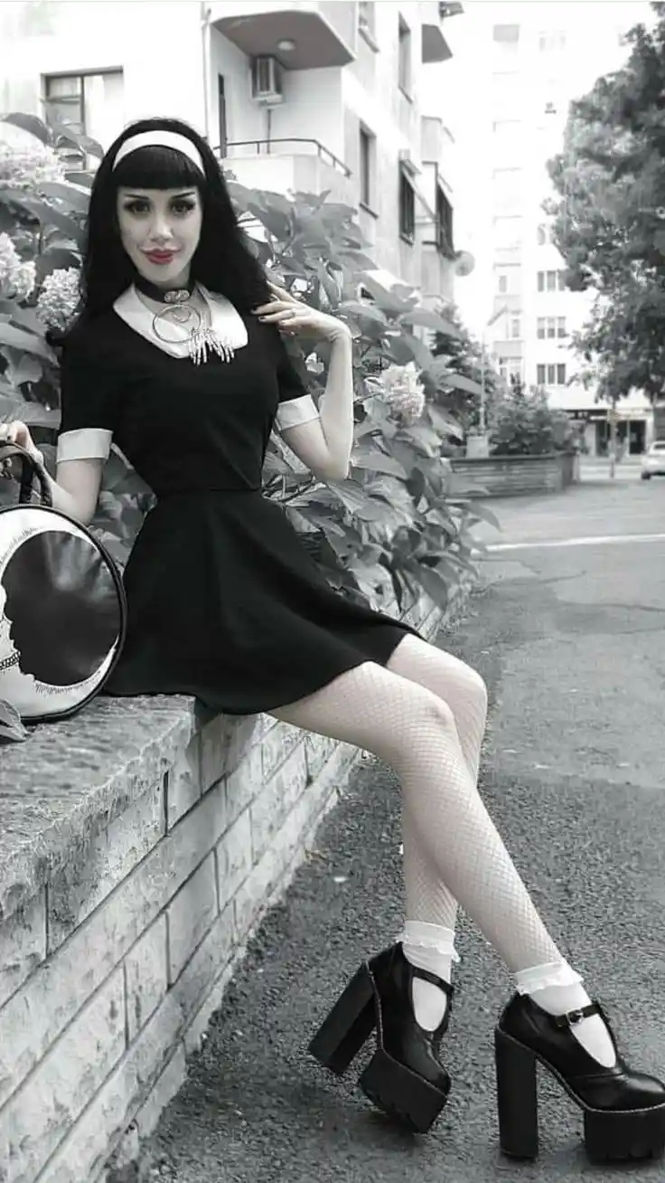 A woman in a black dress with a white collar, fishnet stockings, and platform heels sits on a stone wall outdoors. She wears…