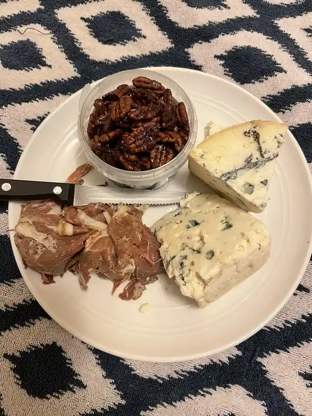 A white plate holds two pieces of blue cheese, a small container of candied pecans, and sliced deli meat, with a serrated…