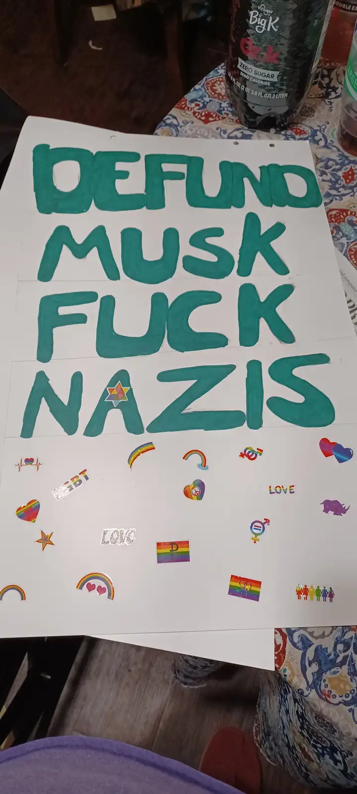 A handmade protest sign with large teal handwritten text reading 'DEFUND MUSK FUCK NAZIS', featuring a rainbow-colored Star…
