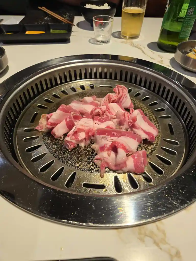 A close-up photo shows raw, marbled beef slices piled on a circular, metal Korean barbecue grill with ventilation slots. In…