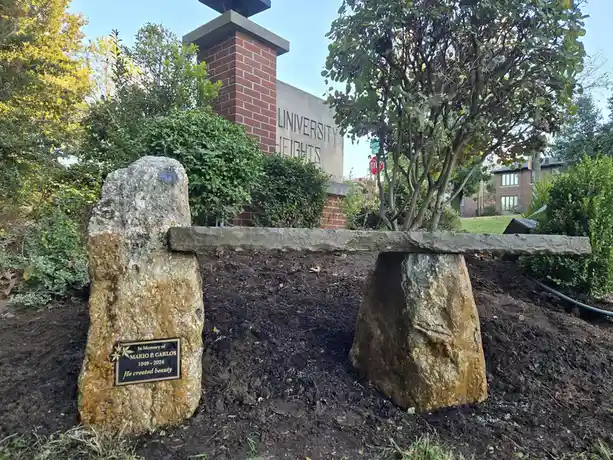 A stone bench made of two large rocks with a horizontal slab, set in a garden with trees and shrubs. In the background, a…