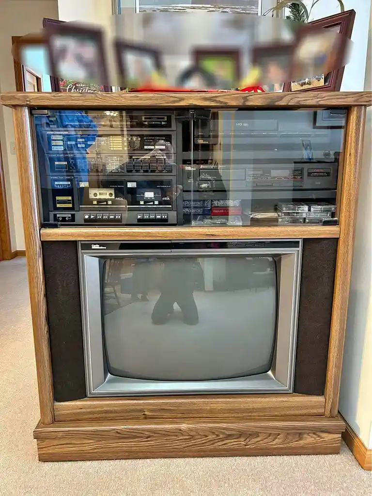 A vintage wooden entertainment center houses a CRT television below a glass-fronted stereo system with cassette decks and…