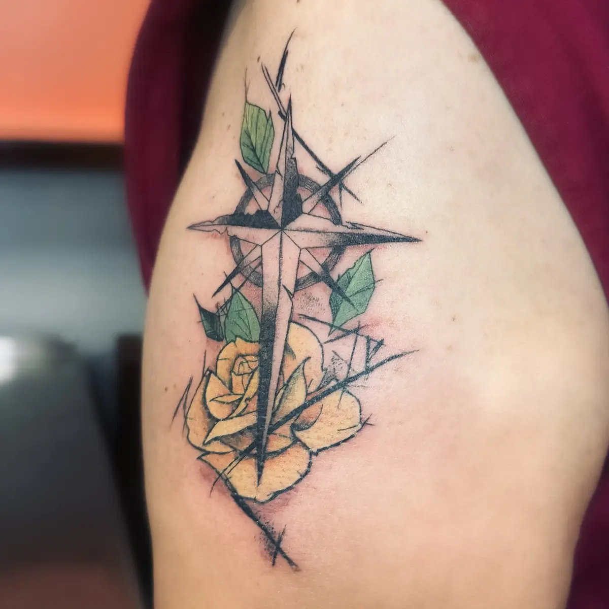A close-up photo shows a fresh tattoo on a person's upper arm, featuring a stylized black compass rose with sharp, angular…
