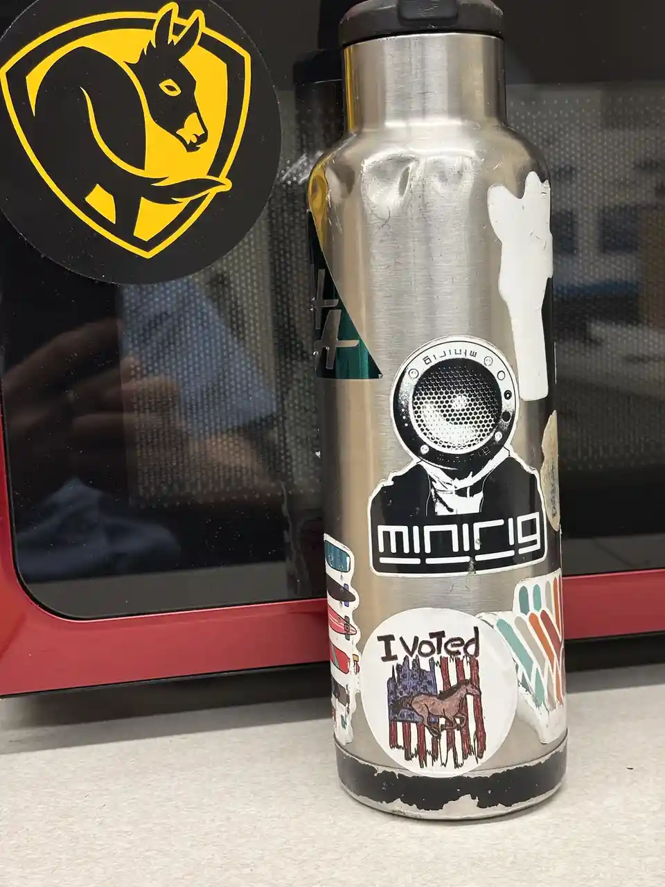 A silver water bottle covered in various stickers, including a prominent circular one that reads 'I Voted' with a horse…