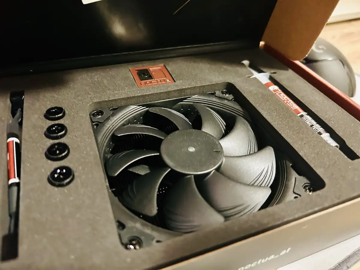 A close-up shot of a Noctua CPU cooler inside its retail box, showing the fan, screws, and included thermal paste tube…