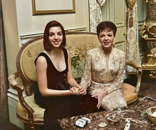 Two women sit side-by-side on an ornate, upholstered couch in a luxurious, vintage room with gold-framed artwork and heavy…