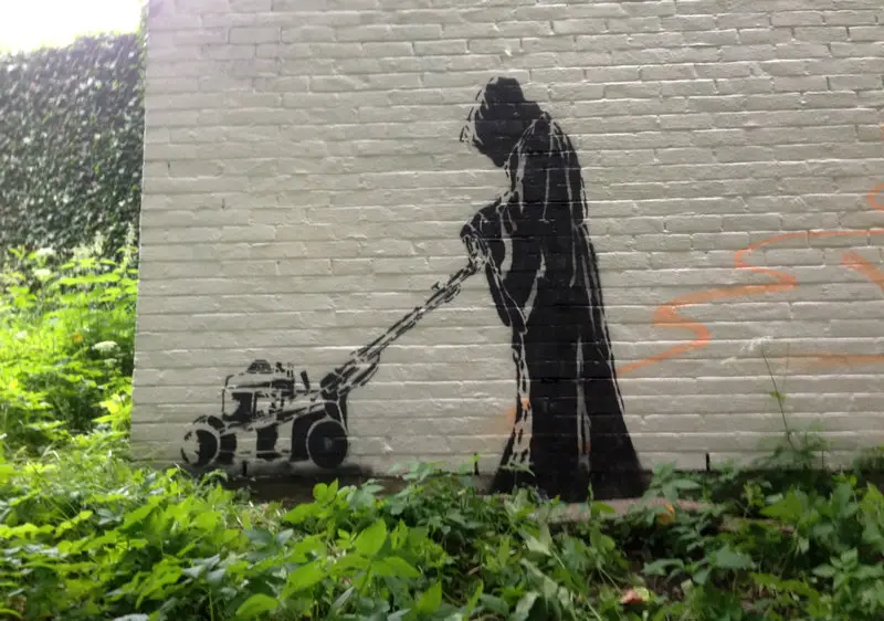 A black stencil graffiti on a white brick wall depicts the grim reaper, wearing a hooded robe, pushing a lawnmower across a…