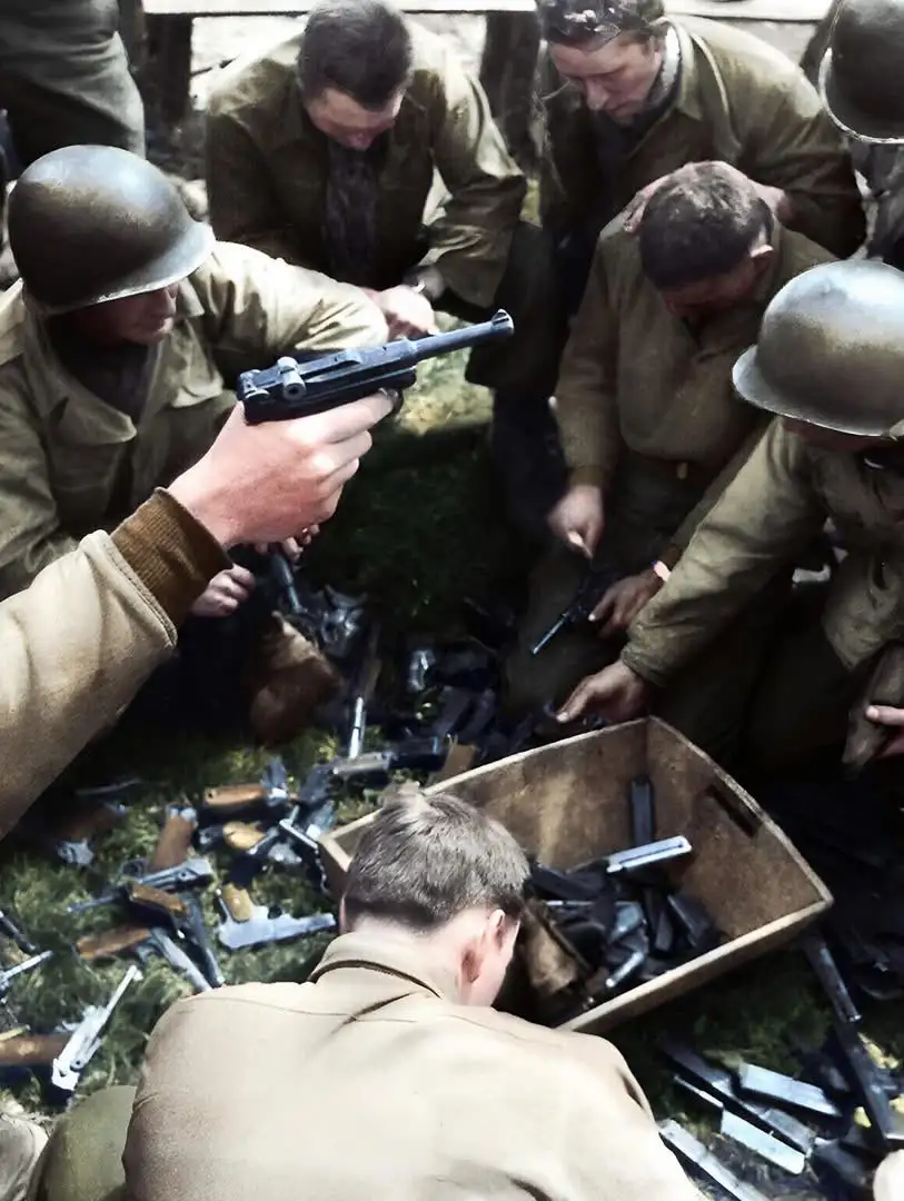 A group of soldiers in military uniforms and helmets are gathered around a pile of firearms on the ground and in a wooden…