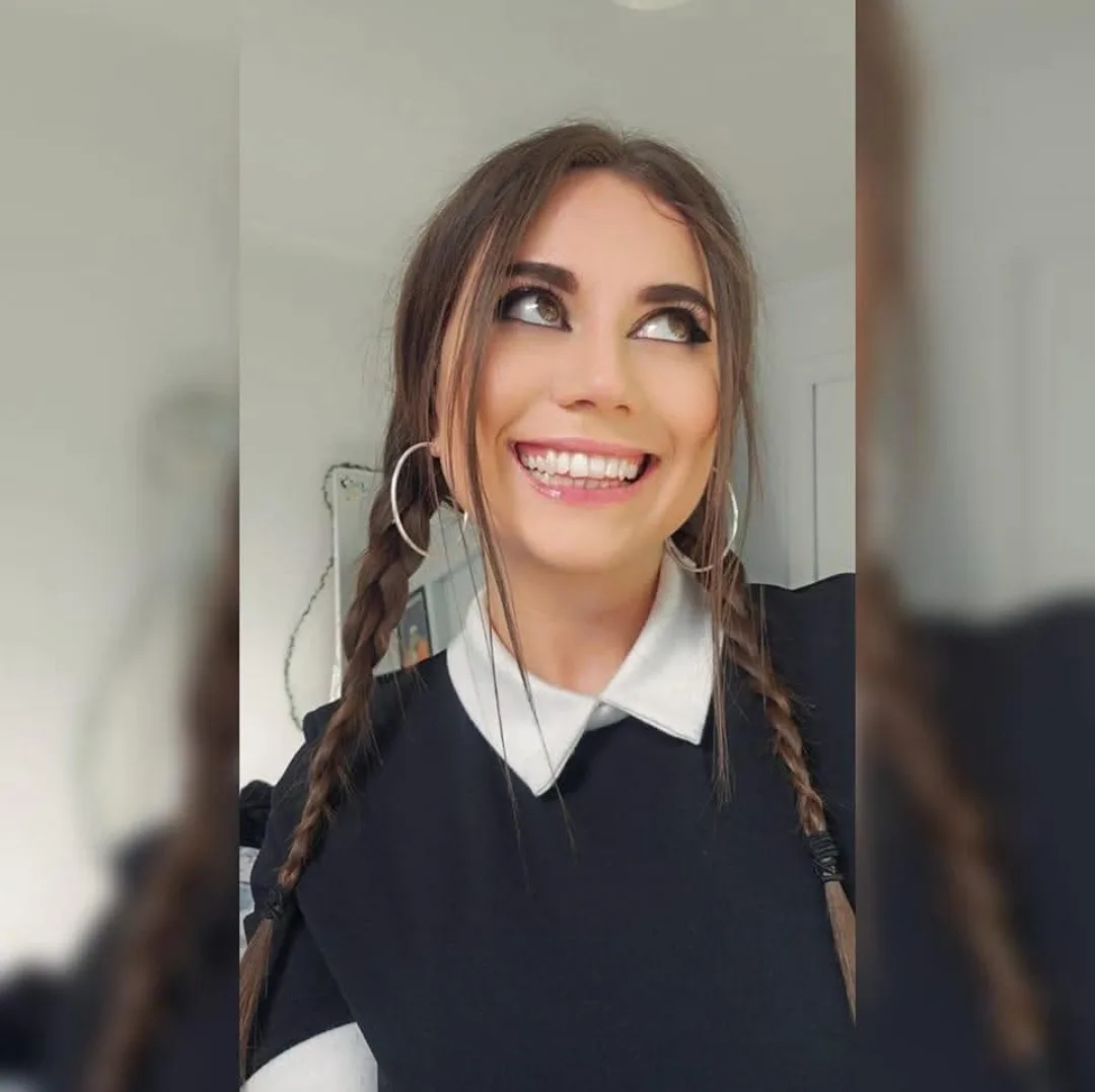 A woman with brown braided hair and large hoop earrings smiles widely while looking upward and to her right. She wears a…