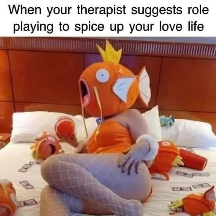 A person wearing a large, orange, cartoonish fish costume with a crown sits on a bed surrounded by smaller fish plush toys…