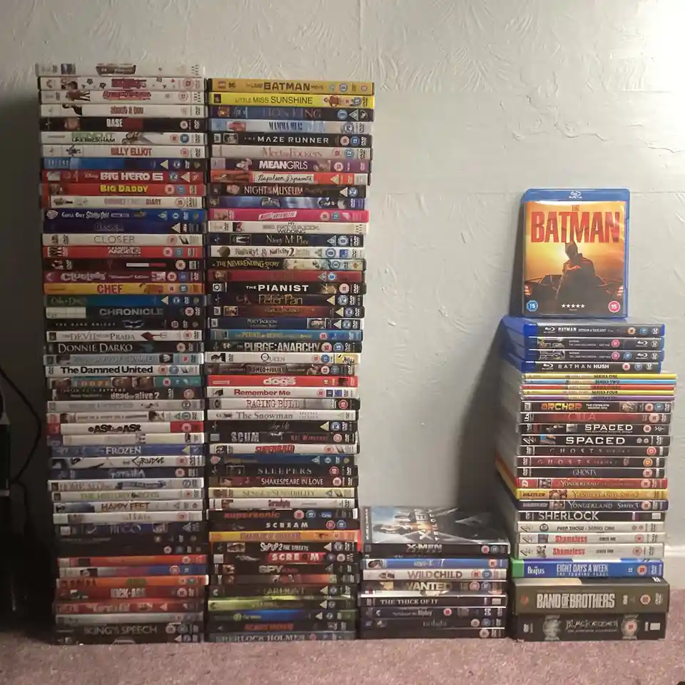 Two tall stacks of DVDs and Blu-rays sit on a carpeted floor against a textured wall, with a single Blu-ray case of 'The…