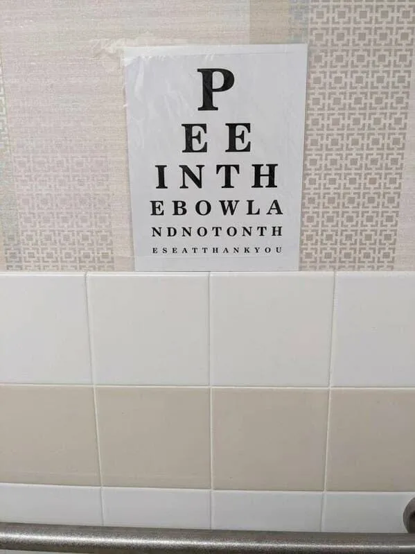 A bathroom wall with white and beige tiles features a sign mimicking an eye chart, but the letters spell out a humorous…