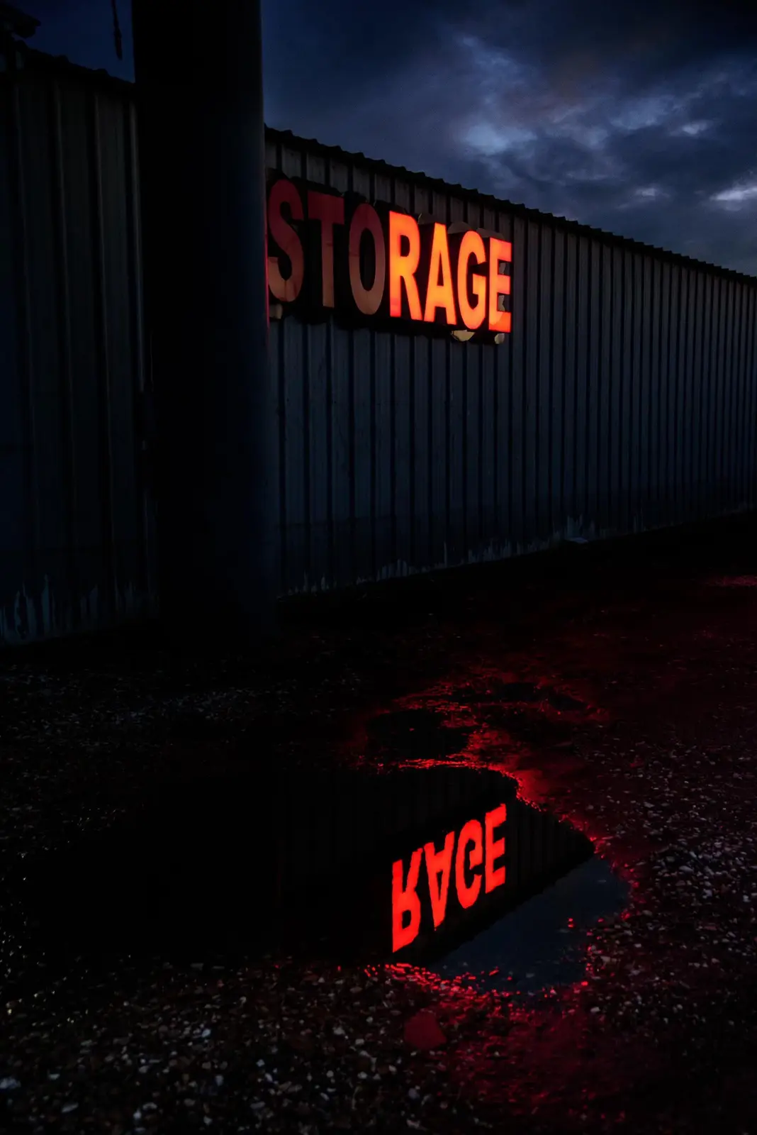 A dimly lit storage facility at night with a glowing red 'STORAGE' sign on a corrugated metal wall. A puddle on the ground…