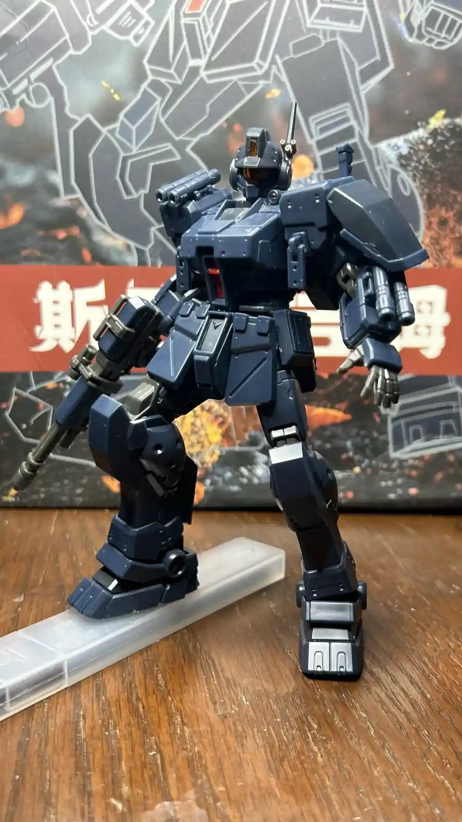 A dark blue, heavily armed robot model stands on a clear plastic stand on a wooden surface, with a backdrop featuring white…