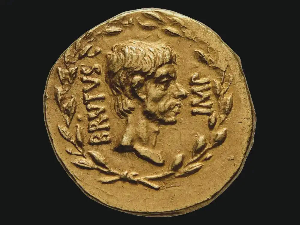 A close-up of an ancient gold coin featuring a profile bust of a man, surrounded by laurel wreath motifs and the Latin…