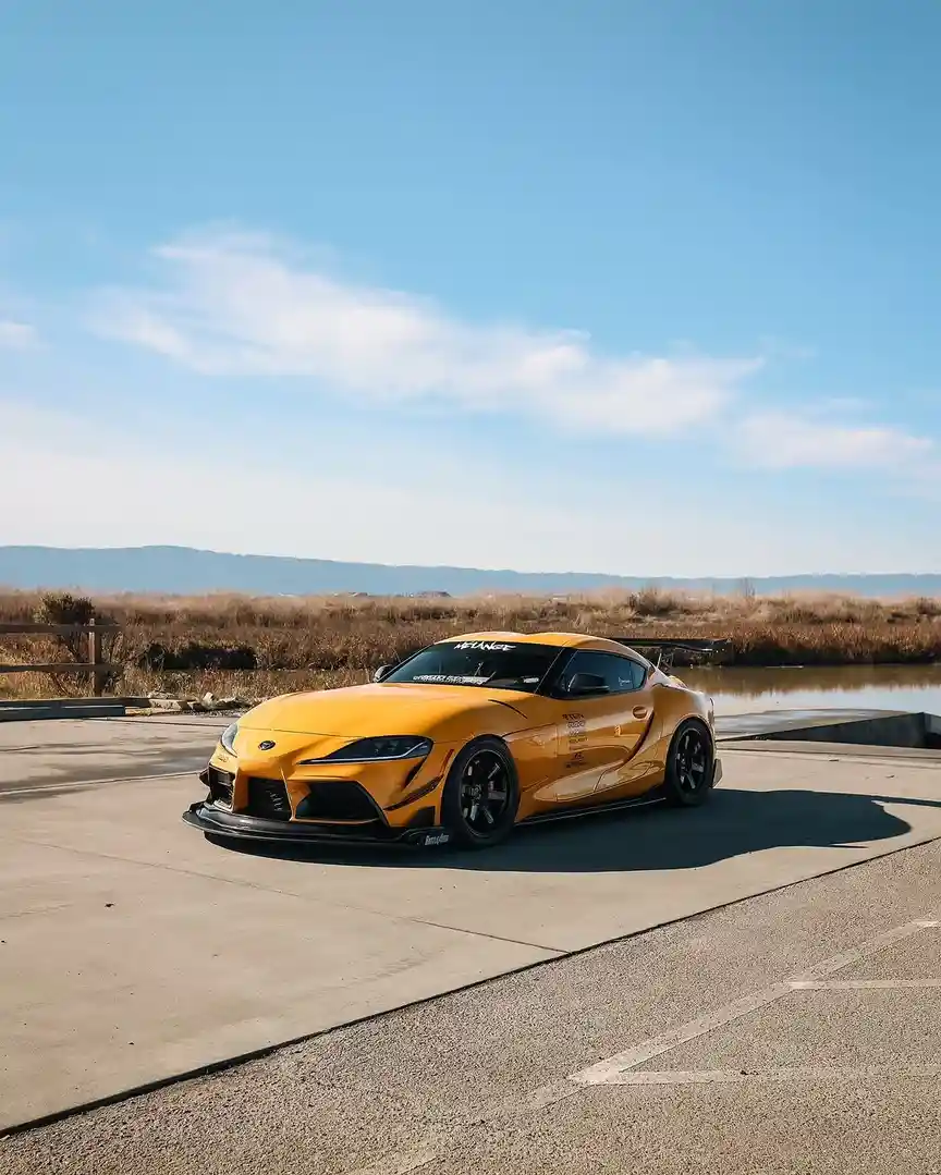 A bright yellow, heavily modified Toyota Supra sports car is parked on a concrete surface near water under a clear blue sky…