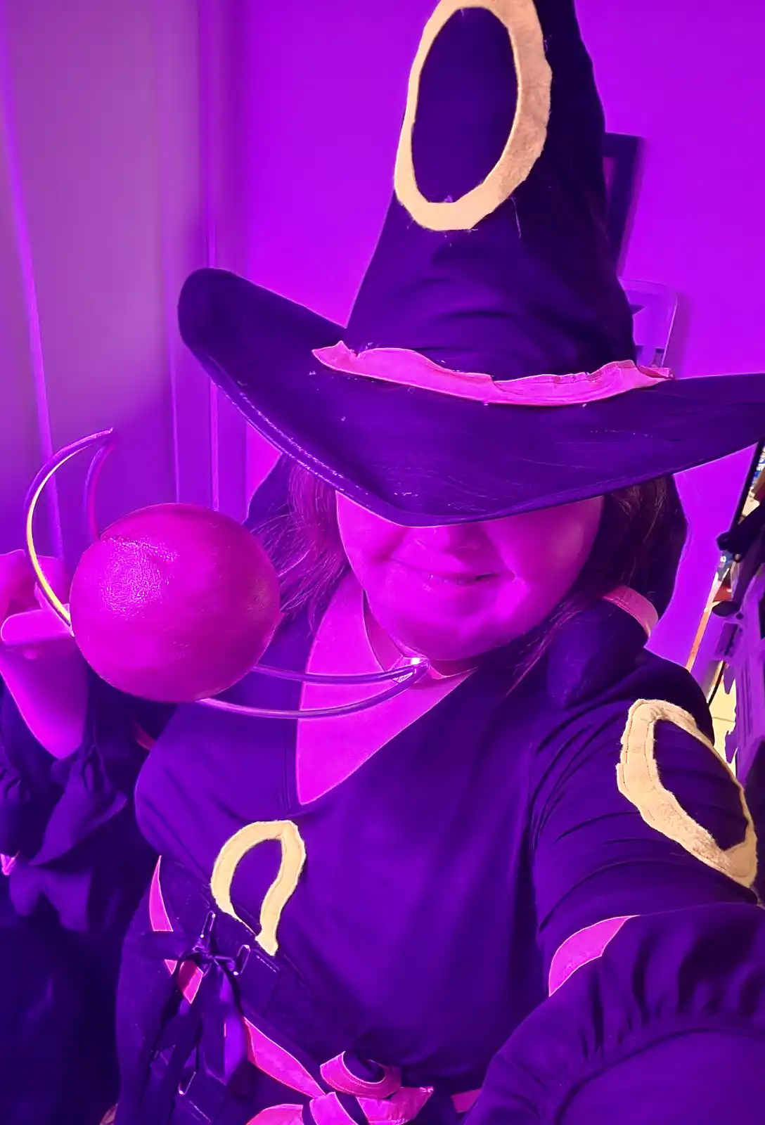 A person in a witch costume with a tall black hat and purple lighting, holding a glowing orb. The costume features gold…